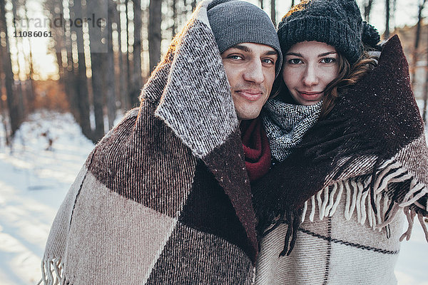Porträt eines lächelnden Paares in Decke gehüllt  während es auf einem schneebedeckten Feld steht.