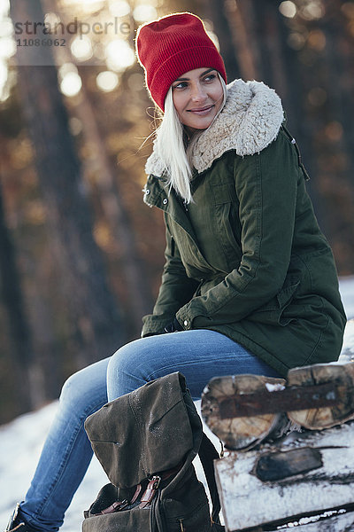 Junge Frau schaut weg  während sie im Winter auf dem Baumstamm sitzt.