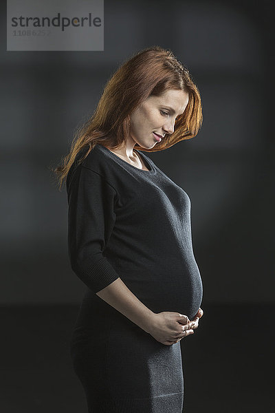 Lächelnde schwangere Frau hält den Bauch vor grauem Hintergrund