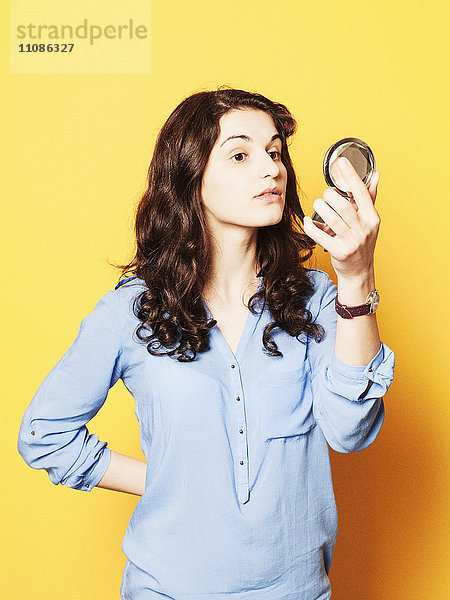 Junge Frau schaut in Puder-Kompaktspiegel vor gelbem Hintergrund