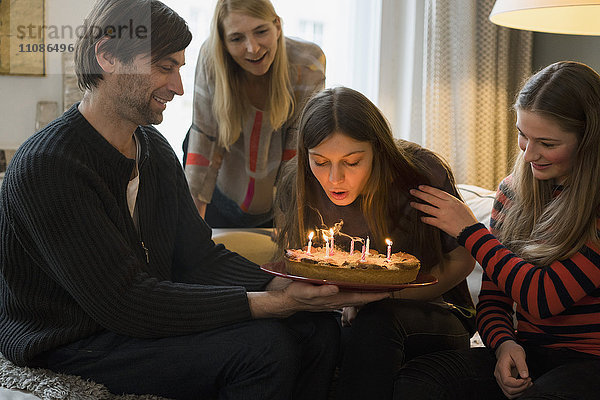 Familie betrachtet junge Frau beim Ausblasen von Geburtstagskerzen zu Hause