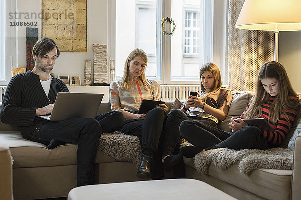 Eltern und Töchter mit Laptop und mobilen Geräten auf dem Sofa im Wohnzimmer