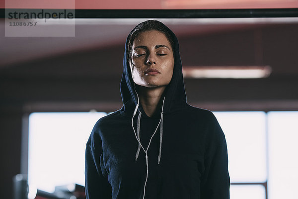 Erschöpfte Frau in Kapuzenshirt stehend mit geschlossenen Augen im Fitnessstudio