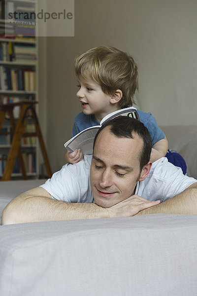 Junge mit Buch sitzend auf dem Rücken des Vaters im Schlafzimmer