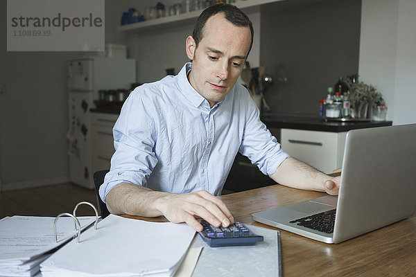 Seriöser Mann  der zu Hause mit Laptop und Taschenrechner über Dokumente arbeitet.