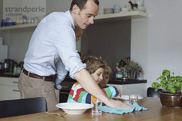 Vater trägt Kleinkind Wischtisch  während Kinder in der Küche essen.