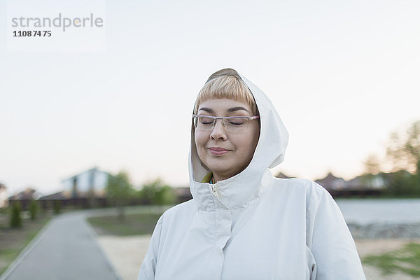 Lächelnde Frau mit geschlossenen Augen mit Kapuze im Park gegen klaren Himmel