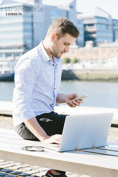 Irland  Dublin  junger Geschäftsmann auf der Bank sitzend mit Laptop und Handy
