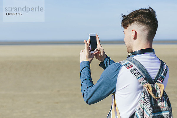 Junger Mann beim Fotografieren mit dem Smartphone am Strand