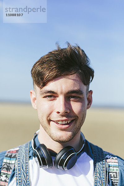 Porträt eines lächelnden jungen Mannes mit Kopfhörer am Strand