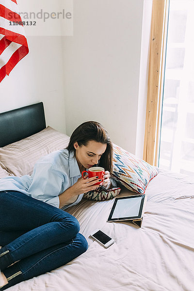 Junge Frau sitzend auf Bett  Kaffeetasse  digitalem Tablett und Smartphone
