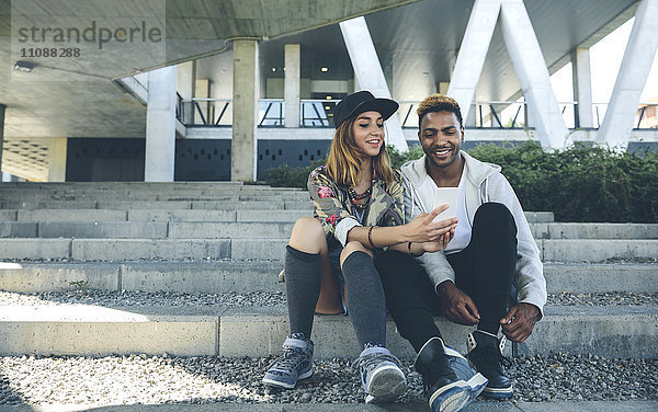 Lächelndes junges Paar sitzt auf einer Treppe und schaut auf das Smartphone.
