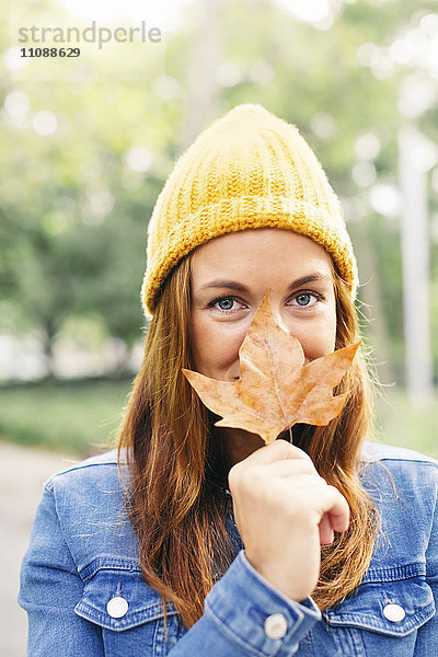 Porträt einer lächelnden jungen Frau mit gelber Mütze  die den Mund mit Herbstblatt bedeckt.