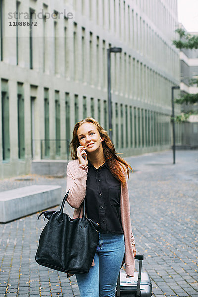 Lächelnde junge Frau mit Rollgepäck und Ledertasche am Telefon