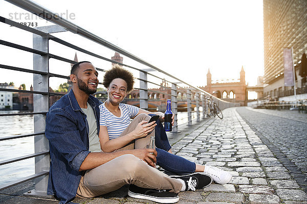 Paar sitzend am Fluss mit Flaschen und Smartphone