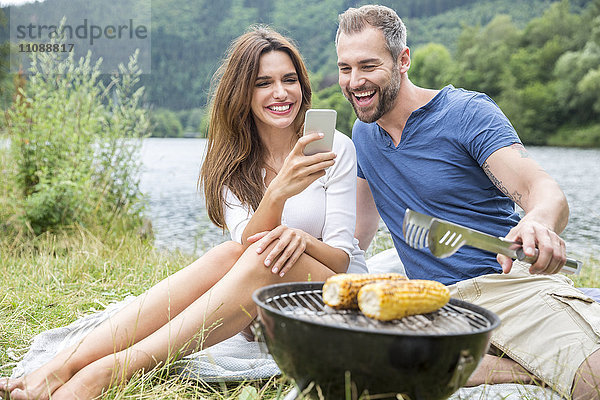 Glückliches Paar mit Handy beim Grillen am Seeufer
