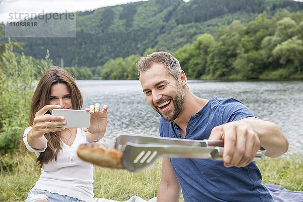 Glückliches Paar macht Handyfoto vom Barbecue am Seeufer