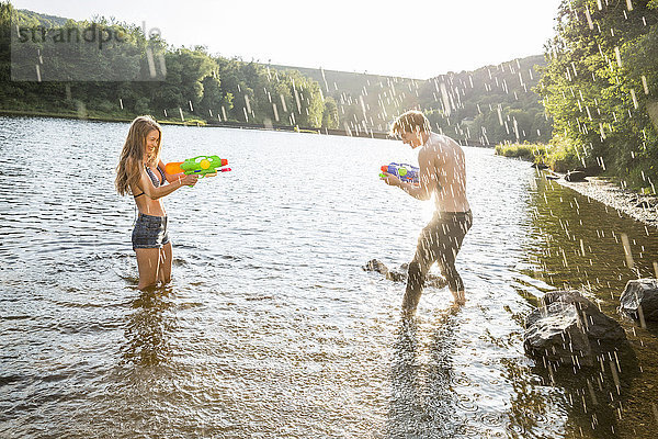 Junges Paar in einem See  das mit Wasserpistolen spritzt.