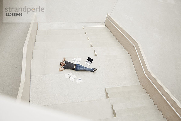 Junger Mann auf der Treppe liegend mit Laptop und Papieren