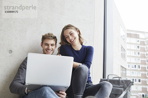 Lächelndes junges Paar sitzt an der Betonwand und teilt sich den Laptop.