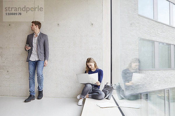 Junge Frau mit Papieren und Laptop und Mann mit Handy an der Betonwand