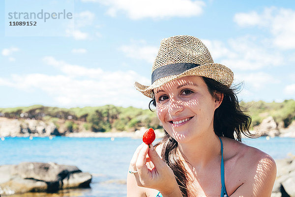 Porträt einer lächelnden Frau mit Strohhut am Strand  die Erdbeere hält.
