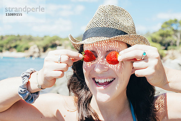 Porträt einer lächelnden Frau mit Strohhut am Strand  die ihre Augen mit Erdbeeren bedeckt.