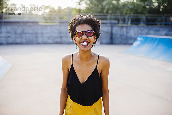 Porträt einer lächelnden jungen Frau in einem Skatepark mit verspiegelter Sonnenbrille