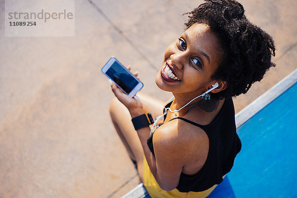 Lächelnde junge Frau mit Kopfhörer und Handy im Skatepark mit Blick auf die Kamera