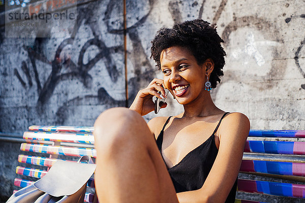Porträt einer glücklichen jungen Frau  die auf einer Bank sitzt und mit dem Handy telefoniert.