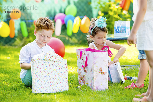 Kleiner Junge und Mädchen beim Auspacken von Geburtstagsgeschenken im Garten