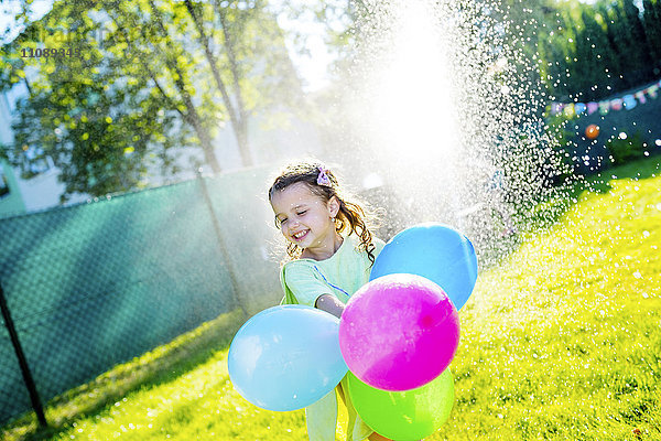 Kleines Mädchen mit Luftballons mit Rasensprenger im Garten