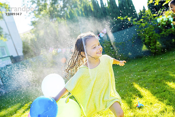 Kleines Mädchen mit Luftballons mit Rasensprenger im Garten
