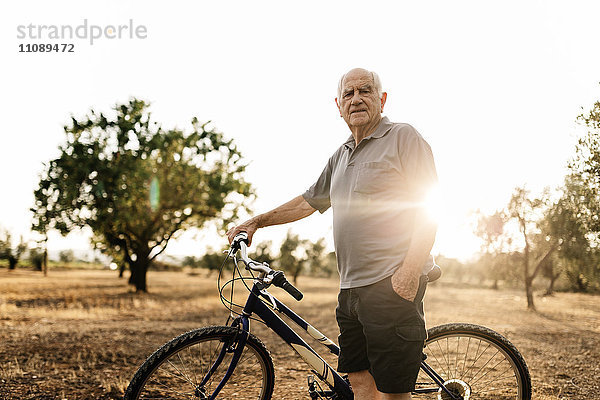Senior mit Fahrrad auf einem Feld in der Abenddämmerung stehend