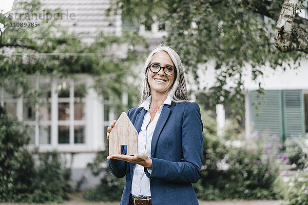 Porträt einer lächelnden Geschäftsfrau mit einem Stück Holz in Form eines Hauses im Garten.
