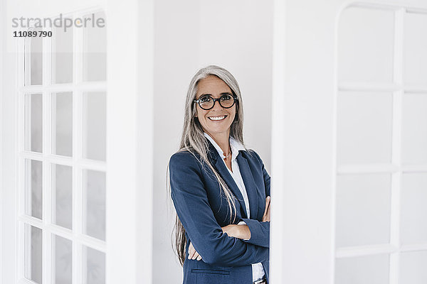 Porträt einer lächelnden Geschäftsfrau mit langen grauen Haaren und Brille zwischen zwei Glastüren.