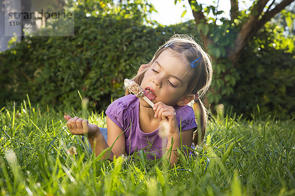 Kleines Mädchen liegt auf der Wiese und isst Eis am Stiel.