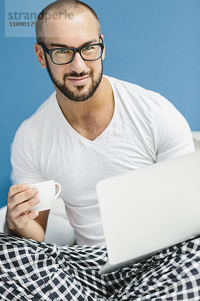Schwuler Mann sitzt auf dem Bett  benutzt Laptop und trinkt Kaffee.