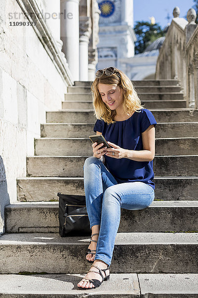 Italien  Udine  lächelnde blonde Frau sitzt auf der Treppe und schaut aufs Handy