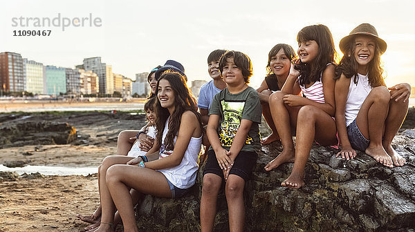 Gruppe von Kindern  die bei Sonnenuntergang auf einem Felsen am Strand sitzen.