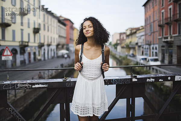 Italien  Mailand  Porträt einer lächelnden jungen Frau mit Rucksack in weißem Sommerkleid auf einer Brücke stehend