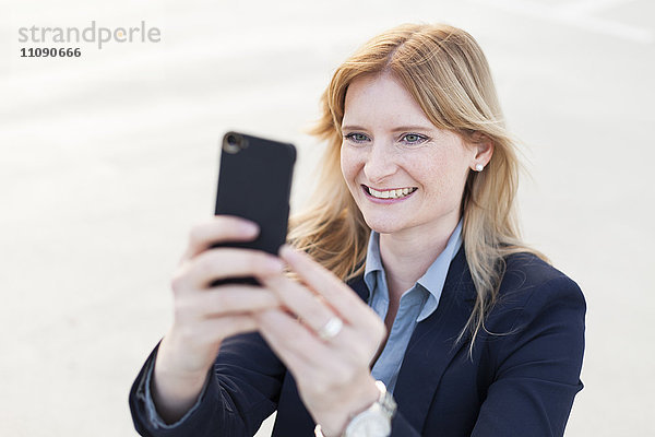 Porträt einer lächelnden  blonden Geschäftsfrau  die sich mit dem Smartphone selbstständig macht.