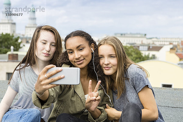 Deutschland  Berlin  drei Teenager-Mädchen sitzen auf dem Dach und nehmen Selfie mit Smartphone.