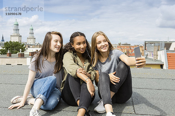 Deutschland  Berlin  drei Teenager-Mädchen sitzen auf dem Dach und nehmen Selfie mit Smartphone.