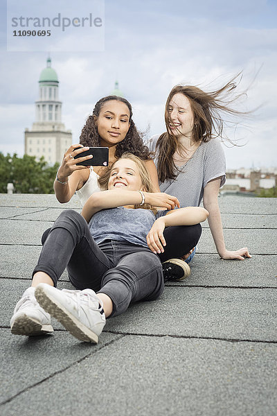 Deutschland  Berlin  drei Freunde sitzen auf dem Dach und nehmen Selfie mit Smartphone