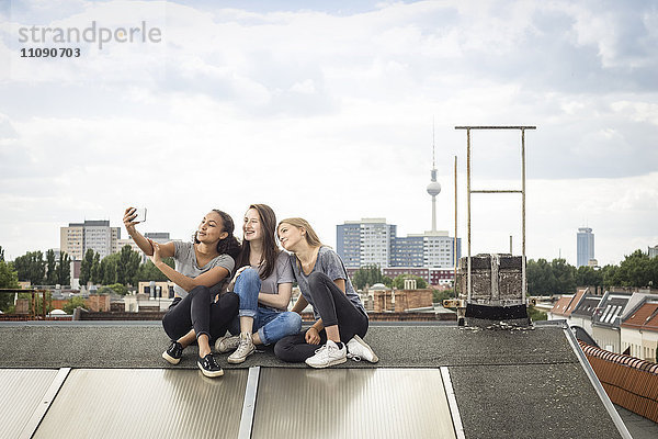 Deutschland  Berlin  drei Freunde sitzen nebeneinander auf dem Dach und nehmen Selfie mit dem Smartphone.