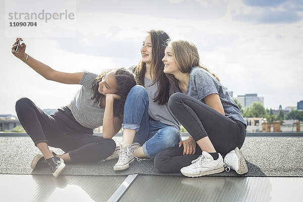 Deutschland  Berlin  drei Freunde sitzen auf dem Dach und nehmen Selfie mit Smartphone