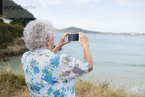 Rückansicht einer älteren Frau  die mit einem Smartphone vom Meer aus fotografiert.