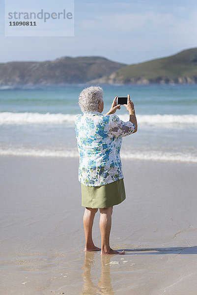 Rückansicht einer älteren Frau am Strand  die mit dem Smartphone fotografiert