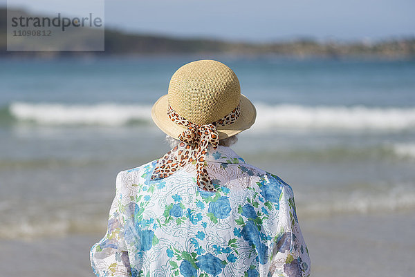 Rückansicht der älteren Frau mit Strohhut am Strand mit Blick in die Ferne